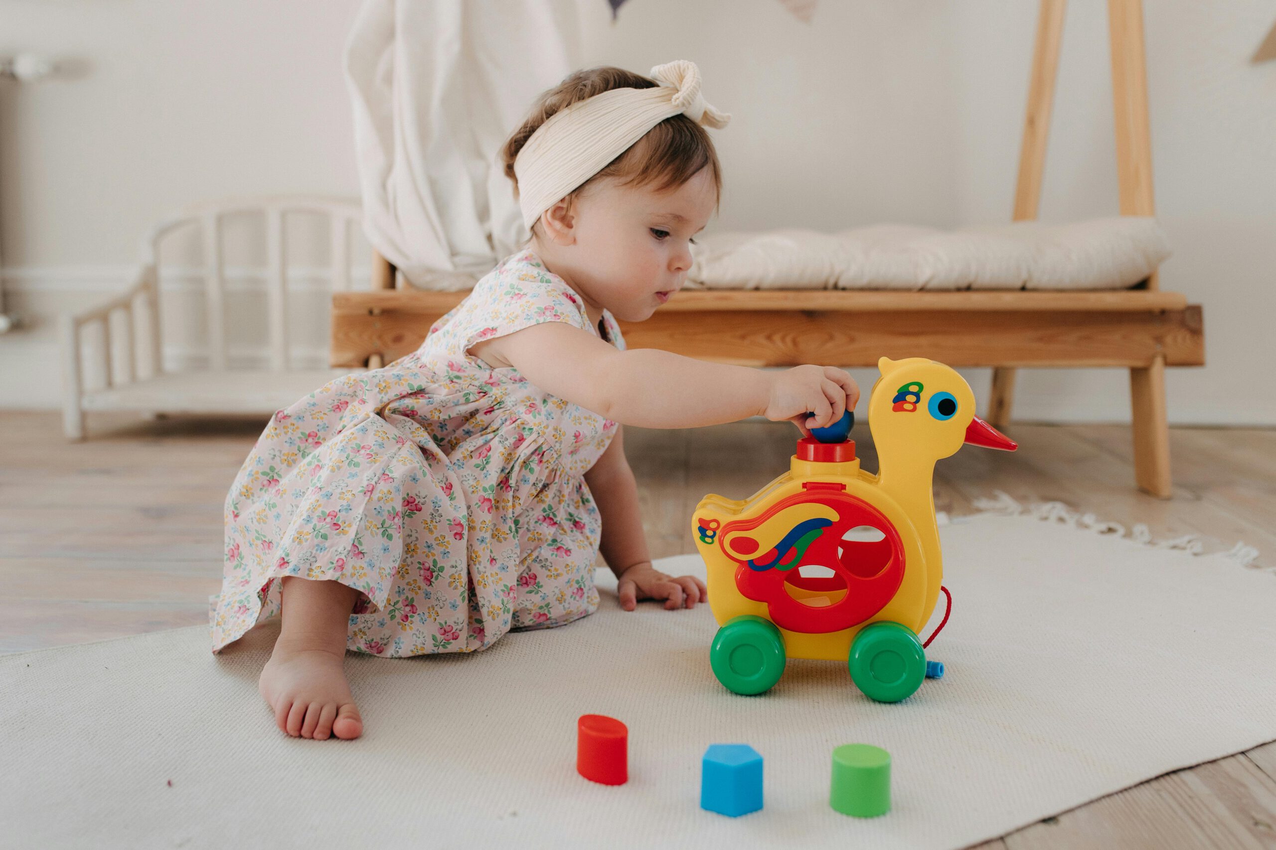 Cute baby girl in a floral dress playing with a colorful duck toy indoors.